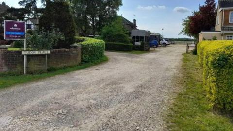 An entrance to the site through a gravelled driveway and bushes on either side. On the left is a street sign that reads "Tudor Place".