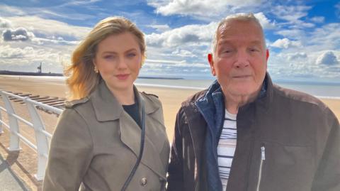 A young woman with blonde hair standing alongside an older man with short grey hair on a promenade along a beachfront. There's golden sand behind them and the sea in the distance. The sky is blue with some clouds. In the left corner of the shot there is a large crane above a harbour. The woman is wearing a grey, buttoned front coat with a leather handbag strap across her front. The man is wearing a dark jacket with a blue top underneath and a white t-shirt. 