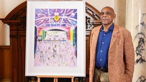 British artist Denzil Forrester - a man smiling, wearing a navy buttoned shirt, a brown tweed suit jacket and light brown trousers, standing to the right of his artwork depicting a reggae festival in Cornwall.