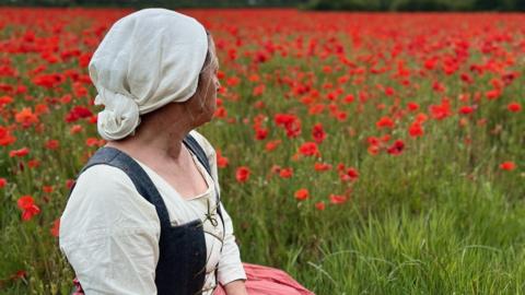 A woman sitting looking out on a field of red poppies, and turned away from the camera. She is in 17th Century dress, comprised of a cream headscarf, plain cream long-sleeved cotton top and a dark grey waistcoat-type garment, fastened together with criss-crossed thread.