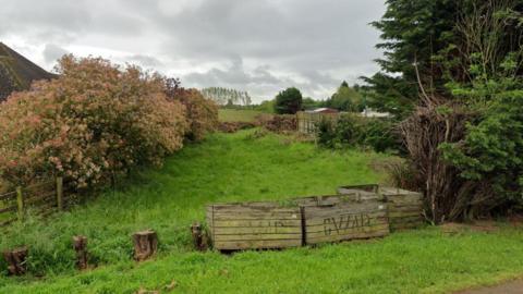 A quiet grassy field, where where 60 homes could be built, is surrounded by dense greenery, including trees and thick bushes. In the foreground are weathered wooden planters. The sky above is heavy with grey clouds.