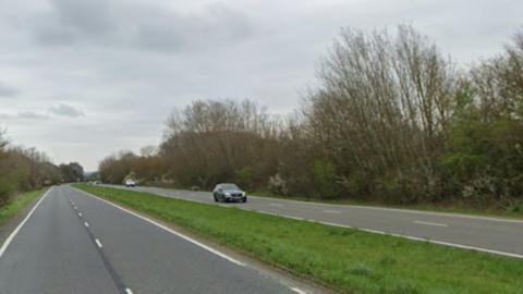 A Google street view image of a dual carriageway - there are two strips of grey tarmac with a green strip of grass between them. There are two grey cars on the right hand carriageway and trees and a grey sky in the background