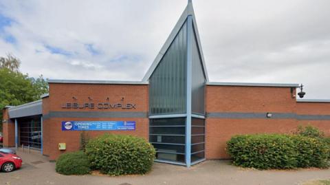 A single storey red brick building with a large vaulted glass wall in the middle. There is a sign that says Leisure Complex on the upper left side of the building and below it is a blue sign that says opening times