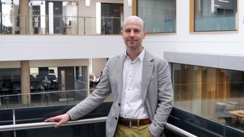 Andrew is wearing a black and white plaid blazer, a white shirt and chinos. He has short hair and is standing with his arm resting on a balcony. He is smiling at the camera. There are offices behind him.