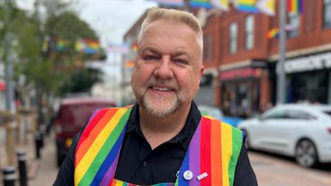 A man is stood in a street. He is wearing a black long sleeved shirt and a rainbow-patterned apron. He has short blond hair and a short grey beard. 