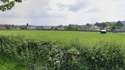 A large green field of a recreation area with a hedge in the foreground and houses beyond it. To the right you can see part of a play park