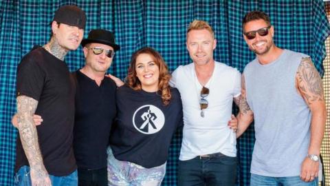 A group of five people posing together in front of a blue checkered curtain. Four men and one woman are smiling at the camera. The men are dressed in T-shirts. jeans and sunglasses, while the woman in the centre is wearing a black sweatshirt and multi-coloured shorts