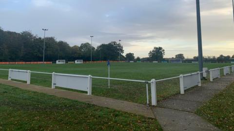 A large, empty football field with white fencing surrounding the edge of the ground. There is a football goal in the distance and two spectator boxes to the left. The sky is cloudy.