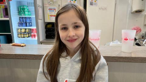 Mia has long brown hair and is wearing a grey hoodie. She is standing in front of a counter at the centre.