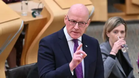 John Swinney, a bald man wearing glasses, a dark jacket, white shirt and purple tie, points a finger to make a point while answering a question in parlieament