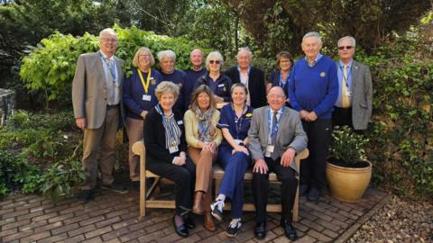 The image shows a group of older adults gathered outdoors in a garden or landscaped courtyard. The scene appears to be taken during daytime, with natural light and leafy greenery suggesting mild weather. The group is posed for a formal or semi-formal group photograph.