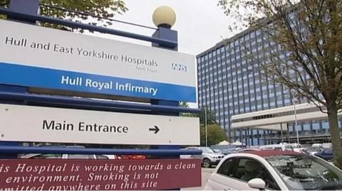 The photo shows the Hull Royal Infirmary with a tall hospital building in the background. In the foreground, a blue and white sign directs visitors to the "Main Entrance" and highlights the hospital’s name. Cars are parked nearby.
