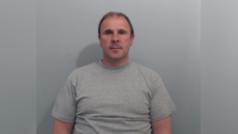 A mug shot of Irvine. It is a head and shoulders image, he is wearing a grey sweatshirt and has short brown hair. 