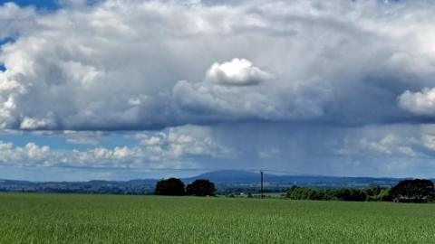 Large cumulonimbus cloud over a grass field with hills in the distance and patchy blue sky above 