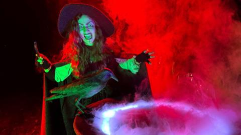 Person dressed as a witch standing next to a cauldron. She is surrounded by fog which is illuminated by red and green light. 