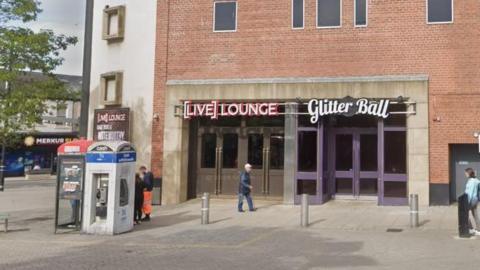 Street view of the outside of the Live lounge. People are walking by the large brown doors in a brick building beneath a sign in white letters with a red border reading Live lounge