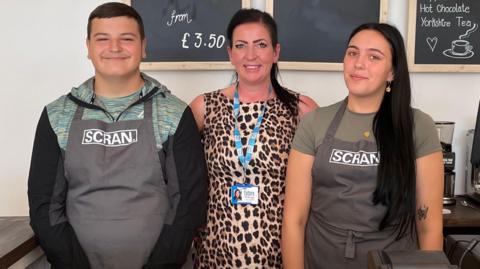 Lauren Meston, the founder of Future Pathways, is stood behind the counter of Scran with 15-year-old Tommy and 17-year-old Freya, who are both wearing branded aprons.