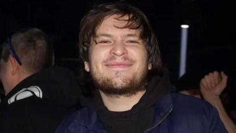 Ezra Tilford-Key in what appears to be a nightclub or party. He is smiling. He has a full beard and moustache, and has medium-length dark hair. He is wearing a black t-shirt and a blue jacket. A light shaft from a spotlight can be seen behind him.