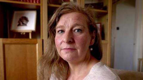 A headshot of Sarah, looking straight at the camera with a serious expression. She is wearing an oat coloured top and earrings with her hair loose. She is sitting in a living room, there are wooden shelves behind her. 