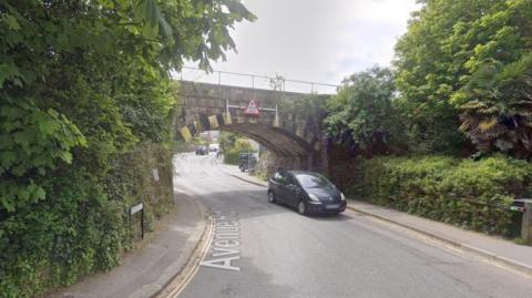 Google Street View image of a black car passing under a railway bridge in Falmouth with green bushes and trees either side.