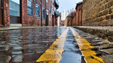 A cobbled road with double yellow lines running down one side. There are buildings on either side of the road. 