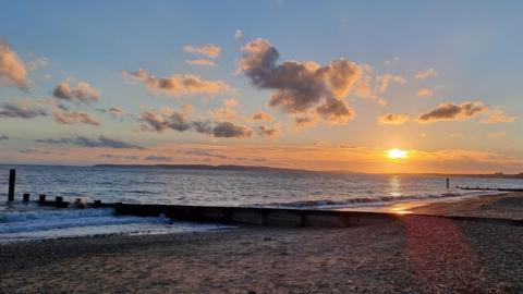 A sky containing clouds contrasting against blue sky, with a low sun sending orange light over a beach and the sea in Lee-on-the-Solent