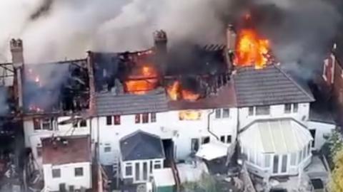 Aerial view of a residential building fire, with flames breaking through the roof of multiple attached houses and dense smoke spreading across the area