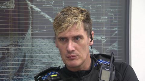 PC Maverick Reddington wearing uniform with police radio attached, sitting in front of a blacked-out office windowk