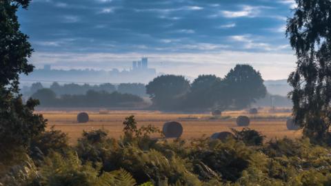 An artistic shot of Lincoln city from a distance over misty countryside. There are hay bales in the foreground and Lincoln Minster can be seen silhouetted in the background. 