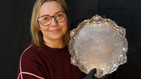 A smiling woman with glasses and a burgundy jumper holds a silver salver aloft. 