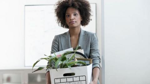 A woman wearing a suit carries a box containing a plant and paper documents