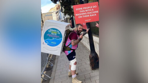 Martin Realey, wearing athletic gear and with a white washing machine strapped to his back, leans against a lamp post and points at a sign on it saying "some people do this with a washing machine on their back". He has short black hair and a trimmed beard, with tattoos on his right forearm.