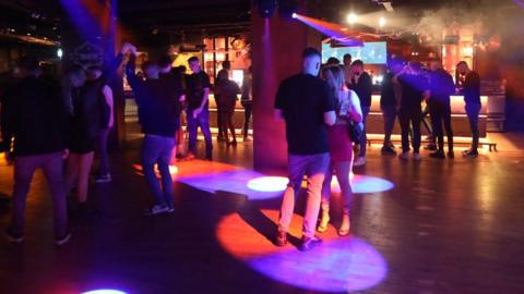 People dancing inside a nightclub. The clubbers are spaced out across the room, some are dancing and some are talking.  Multi-coloured lights are lighting up the dark room.