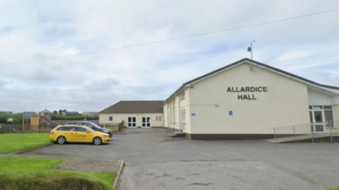 A cream coloured building with multiple doors, surrounded by a concrete car park and grass. There is a yellow and a black car to the left of the building, and the building has the words 'Allardice Hall' on the front of it.
