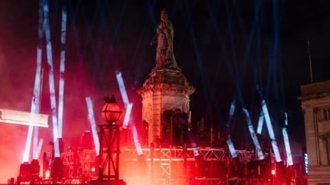 A statue is illuminated in Queen Victoria Square at night time. The large stone statue stands on the top of a large plinth and is surrounded by beams of white and red light.