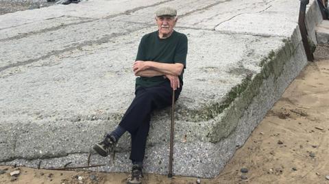 An elderly man in a dark green t-shirt and black trousers sitting on a large slab of grey rock. He is wearing a flat cap and is holding a brown walking stick. There is sand surrounding the rock