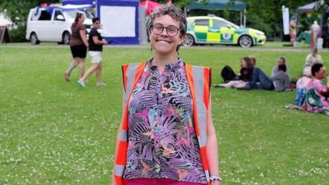 A woman stood on green grass. She is wearing a brightly coloured top, an orange high visibility vest and pink trousers. Behind her the grass is green and full of white flowers. There are people in the background sitting on the grass. 