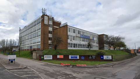 The large building stands on slightly raised ground on a corner with business names on signs below on a grass verge. The building straddles the corner plot.
