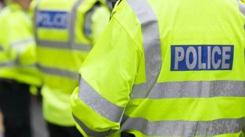 Three police officer are standing next to each other. They are all wearing yellow hi-vis jackets with silver stripes, with police written on the back in silver. 