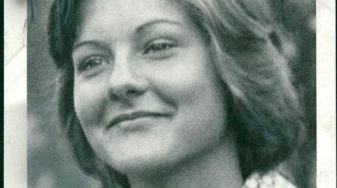 A black and white photo of a smiling young woman looking away from the camera. She is wearing a 1970s-style shirt with a geometric pattern and big wing collars.