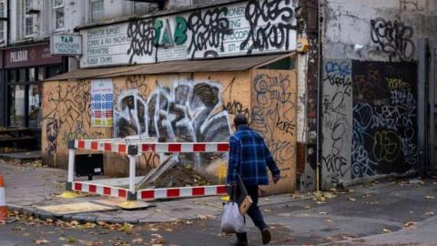 A woman walks past a boarded up shop covered in graffiti