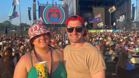 A woman and a man stand illuminated by sun in front of a festival stage, with a large crowd watching the stage.