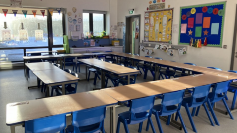 Image shows an empty primary school classroom, with small blue chairs lined up around several pushed-together desks. 