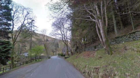 The narrow road which winds into the distance. There are trees on both sides of the road, and a former chapel on the right, with a hill in the distance.