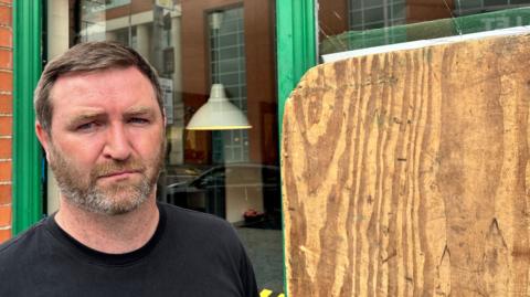 Gareth McKinstry, with short dark hair and a black t-shirt, standing in front of his chip shop