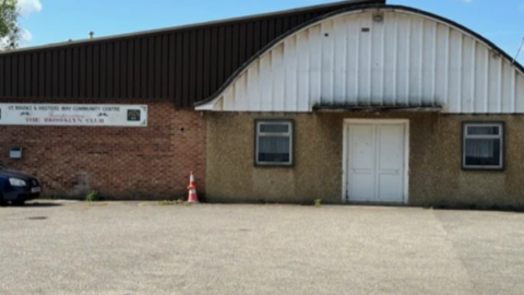 The centre, when it closed, with a dated facade 1940s facade of concrete and brick with white entrance door and a white arch-shaped roof above. A blurred sign on the left still has the former name St Marks and Hesters Way Community Centre on it 