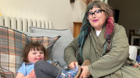 A woman with pink and green dreadlocks smiles into camera perched on a  sofa next to her four-year-old daughter who has Down's syndrome.