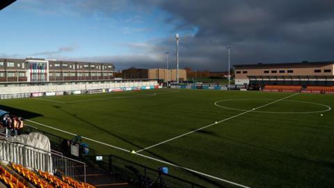 Arbour Park, with a football pitch in the centre of the picture, a partly-seated stand on the far side of it and a terrace behind the goal, with a building behind that 