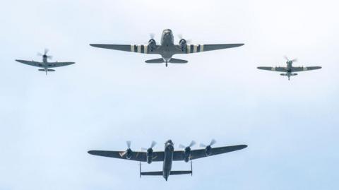 Silhouettes of four aircraft, two large two small on a pale blue sky. The largest aircraft has four rotors.