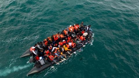 A dinghy pictured from above by a drone. The small boat is filled with people wearing fluorescent life jackets 